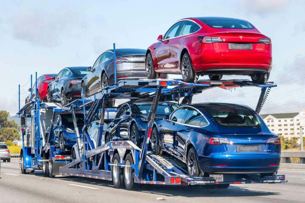 Blue Open Trailer with 8 Tesla Model 3s traveling away from the photo.