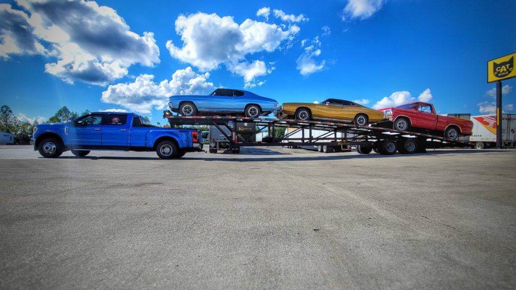 A blue dually pickup truck with a 3-car wedge trailer hauling three classic cars. Open and enclosed car shipping service.