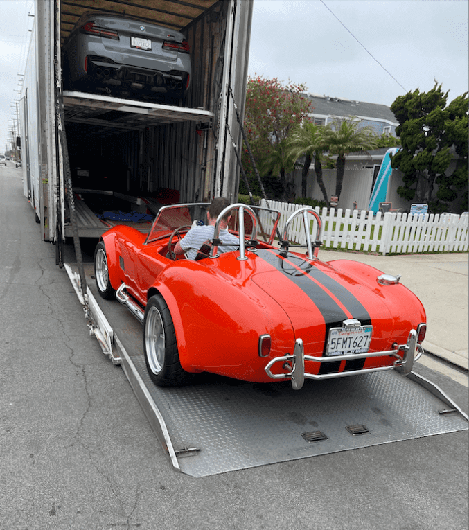 A 1960s Shelby Cobra in orange with black racing stripes getting loaded into an enclosed auto transport trailer.