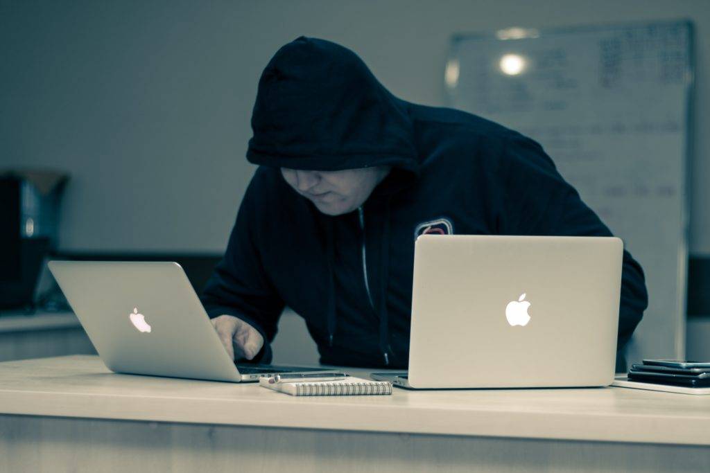 Man in a black hoodie using two laptops. A legitimate company will have a shipping agent with excellent customer service who will explain the common vehicle transport scams to you.