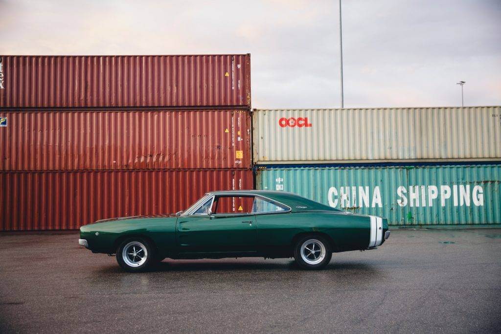 A green 1970 Dodge Charger in front of 40ft containers at an Ocean port. 