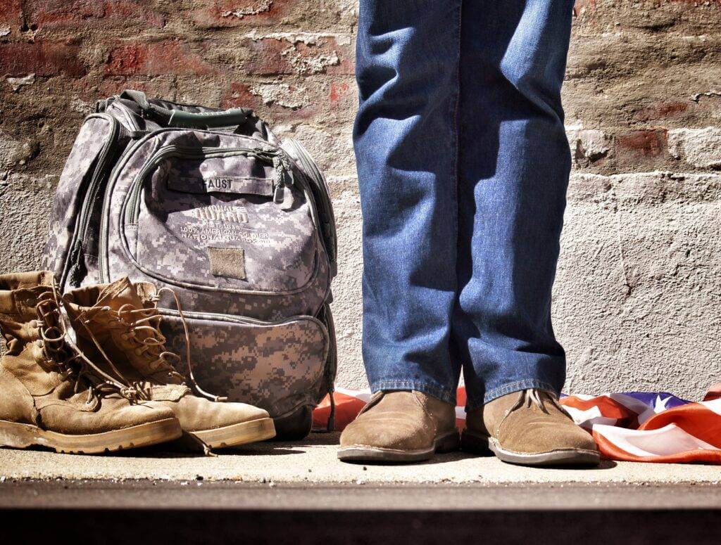 A view of someone's legs from the knee down. They are wearing blue denim jeans and light brown shoes. To their left are a pair of military boots and an army backpack. Make sure to inquire about military pcs vehicle shipping. Many military ship their privately owned vehicle with door to door service.