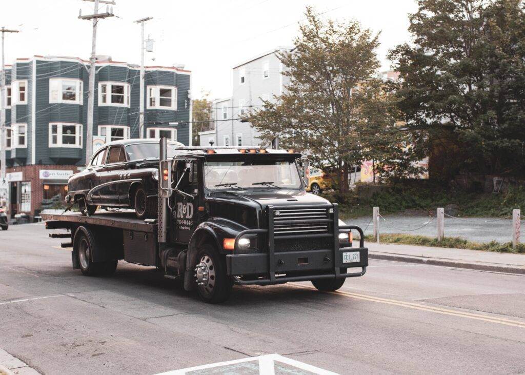 A black flatbed tow truck with a classic car on the bed. Long-distance car towing is our specialty. At National Transport Services, our auto transport services are second to none. We provide top-quality services and the best long-distance towing rates.